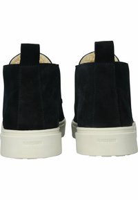 Botas de ante negras hasta el tobillo con suelas de goma blanca y forro de piel de color claro. Presentan una lengüeta en la parte trasera para facilitar su uso.