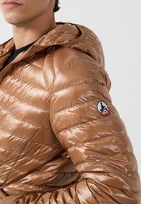 Giacca a vento marrone con finitura lucida, caratterizzata da cuciture orizzontali, cappuccio e una toppa con il logo sulla manica sinistra.