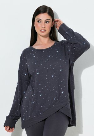 Kvinde med mørkt hår iført en løs mørk sweater med et lyseblåt stjernemønster og matchende mørke leggings, poserer med den ene hånd nær øret.