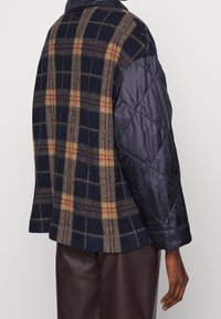 Quiltad marinblå nylonärm och krage i kombination med ett rutigt ullmaterial i mörkblått, beige och burgundy. Avslappnad passform och distinkta texturer.
