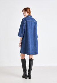 Vestido de mezclilla con cuello, mangas tres cuartos y un corte holgado. Color azul oscuro con detalles de costura visibles. Botas negras hasta la rodilla.