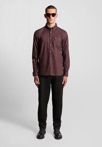 Camicia button-up bordeaux con taschino sul petto, abbinata a pantaloni neri texturizzati e stivali neri con lacci. Modello che indossa occhiali da sole scuri.