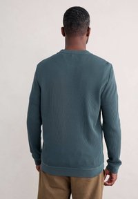 Maglione lungo a maniche con tessuto testurizzato e costine di colore verde scuro, colletto tondo e polsini aderenti, visto da dietro.