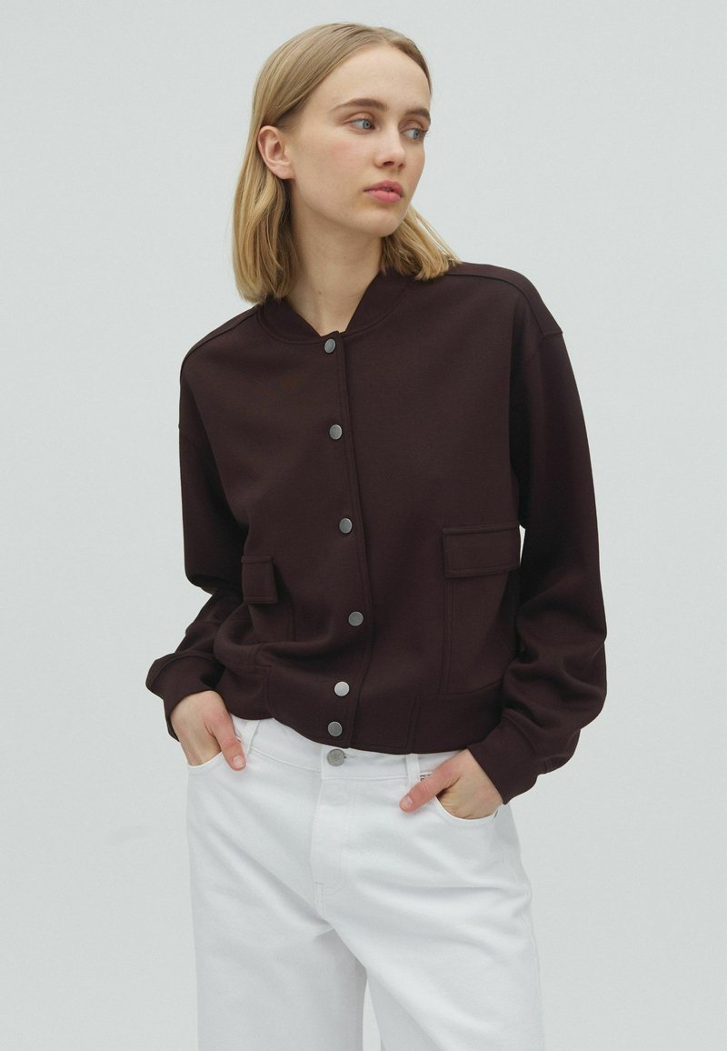 Jeune femme aux cheveux blonds portant une veste boutonnée marron foncé et un pantalon blanc, les mains dans les poches, regardant sur le côté.