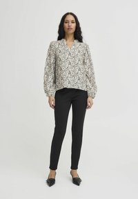 Bluse mit floralem Muster in Creme mit schwarzen Akzenten, V-Ausschnitt und Puffärmeln, kombiniert mit schwarzen Slim-Fit-Hosen und spitzen Schuhen.
