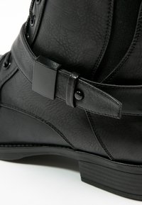 Stivaletto sintetico nero alla caviglia con superficie texturizzata, una fascia larga con fibbia quadrata e tacco impilato.