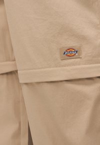 Μπεζ παντελόνι Dickies με κοντινό πλάνο στη μικρή ορθογώνια ετικέτα λογότυπου ραμμένη κοντά στη ραφή του υφάσματος.