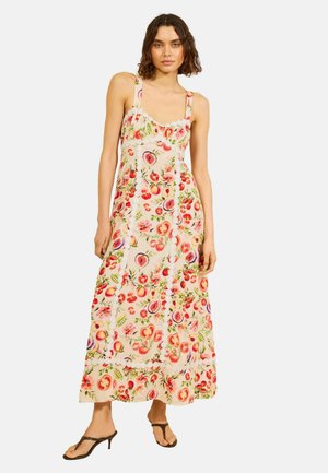 Kvinde iført en ærmeløs, ankel-lang kjole med frugt- og blomsterprint, kombineret med sorte hælhælede sandaler.