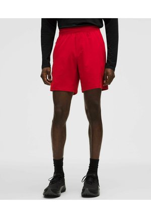 Mand iført lyse røde atletikshorts, sort langærmet trøje, sorte sokker og sorte løbesko, stående mod en ensartet lys baggrund.