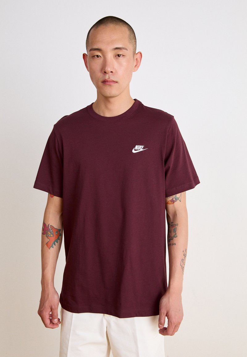 Burgundin värinen puuvillainen t-paita, jossa on valkoinen Nike-logo. Lyhyet hihat, pyöreä kaula-aukko ja rento malli. Suora rakenne ilman kuvioita.