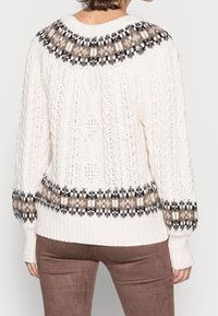 Femme portant un pull crème à torsades avec un empiècement et des poignets à motif noir, marron et beige, associée à un pantalon marron, vue de dos.