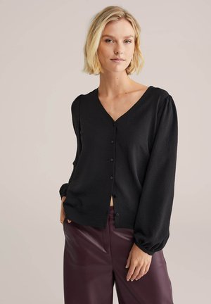 Schwarze V-Ausschnitt Strickjacke mit strukturiertem Finish, langen Puffärmeln und vorderem Knopfverschluss; kombiniert mit figurbetonten dunklen burgunderfarbenen Hosen.
