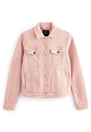 Femme marchant devant un fond orange, portant une veste rose clair, un pull blanc, un jean bleu foncé et des baskets blanches.