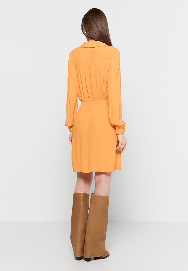 Pomarańczowa sukienka koszulowa z długimi rękawami, kołnierzykiem, zwężoną talią i gładką fakturą. W połączeniu z brązowymi wysokimi botkami.