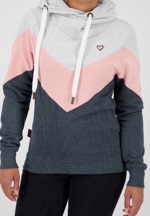 Kobieta w bluzie z kapturem w kolorach szarym, różowym i ciemnoniebieskim z białymi troczkami, z małym emblematem serca na piersi.