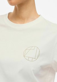 Camiseta de algodón blanca con un sutil logo geométrico dorado en el área del pecho izquierdo; cuello redondo y textura suave.