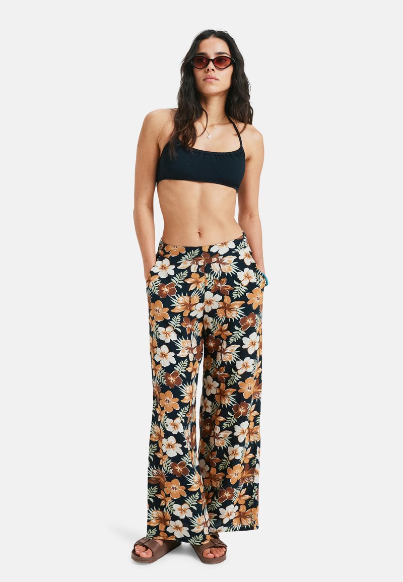Floral wide-leg pants with a black base, featuring white and orange flowers, and side pockets; paired with a dark bikini top and sandals.