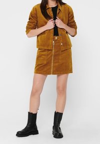 Set di giacca e gonna in corduroy giallo senape, con chiusura a zip, tasche e abbinato a stivali neri alla caviglia. Texture liscia.