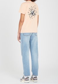 Perzikkleurig t-shirt met een achterafbeelding van een spinnenweb en een diamantvorm. Gecombineerd met lichtblauwe, loszittende jeans en sneakers.