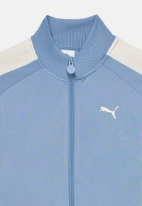 Lys blå zip-jakke med ribbestrikket krage, fargeblokkert design og hvit Puma-logo på brystet, med jevn stoffstruktur.