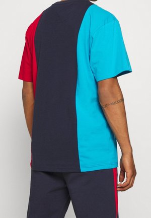 Mann trägt kurzärmeliges T-Shirt mit vertikalen Paneelen in Rot, Navy und leuchtendem Blau, kombiniert mit navyblauen Hosen mit rotem Streifen.