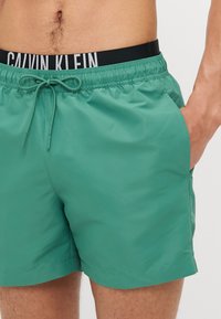 Șorturi de înot verzi, fabricate dintr-un material ușor și neted. Au o talie elastică cu un logo Calvin Klein alb și negru și un șnur de ajustare.