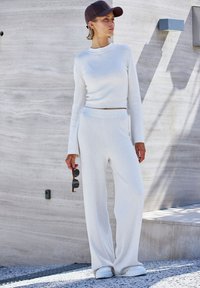 Baltas megztas ilgarankovinis viršus, derinamas su plataus pjūvio kelnėmis. Modelis laiko saulės akinius ir dėvi tamsią kepurę. Tekstūruota medžiaga.