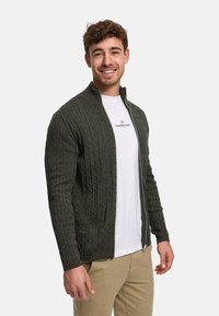 M örkgrön cardigan med dragkedja i stickad struktur, med ett texturerat kabelmönster, ståkrage och ribbade muddar, lager-på-lager över en vit t-shirt.