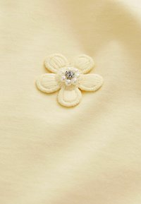 Petite fleur en tissu crème avec cinq pétales brodée sur un tissu jaune pâle, centre décoré de petites perles et d'une pierre claire.