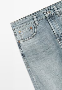 Lichtblauwe spijkerbroek met voorvak, muntvak met klinknagels, riemlussen en knoopsluiting op een witte achtergrond.