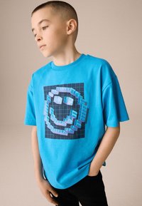 Turquoise T-shirt met korte mouwen en een gepixeld smiley-gezicht op een zwarte rasterachtergrond. Gemaakt van zacht katoenen stof.