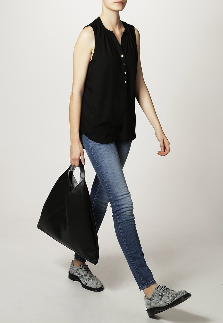 Femme portant un chemisier noir sans manches, jean skinny bleu, chaussures à motif peau de serpent, tenant un grand sac cabas noir, marchant devant un fond uni.