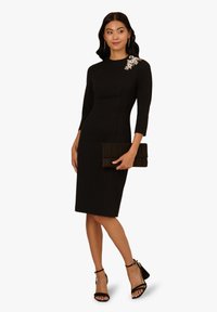 Robe noire ajustée avec des manches trois-quarts, embellissement décoratif sur une épaule, associée à une pochette noire et des talons.