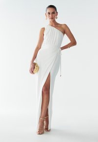 Vit enaxlad klänning med rynkad överdel, sidospalt och knytning i midjan. Kompletteras med guldsmycken och remsandaler.