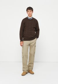 Giovane uomo in piedi con le mani in tasca, indossando un maglione di maglia a treccia marrone, una camicia blu, pantaloni cargo beige e scarpe beige su sfondo bianco.