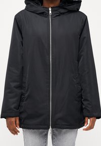 Černá kapucňová bunda se zipem vpředu, postranními kapsami a fleecovou podšívkou viditelnou na manžetách a kapuci, nošená přes světle šedé džíny.