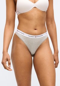 Pelēkas kokvilnas stringa biksītes ar baltu jostasvietu, uz kuras ir melnā, sarkanā un zilā krāsā rakstīts "TOMMY HILFIGER" logotips. Gluda tekstūra un pieguļošs dizains.