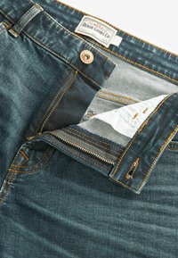 Blugi din denim albastru, cu fermoar, buton din cupru, cusături portocalii contrastante și un eticheta "Denim Goods Co" în interiorul taliei.