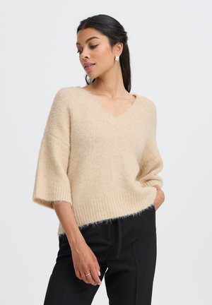 Femme aux longs cheveux foncés porte un pull beige à col en V avec de larges manches et un pantalon noir, debout avec une main dans la poche devant un fond uni.