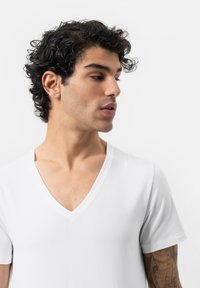 Jonge man met krullend donker haar en een getatoeëerde arm die naar links kijkt, gekleed in een eenvoudige witte V-hals T-shirt tegen een witte achtergrond.