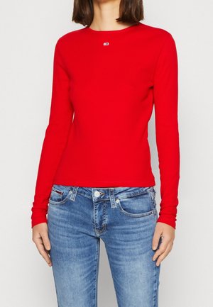 Person trägt ein schlichtes rotes Langarmshirt und blaue Jeans und steht vor einem neutralen Hintergrund.