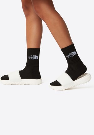 Hvite sandaler med svart stropp, kombinert med svarte rillede sokker som har en hvit logo. Glatt tekstur med en polstret såle.