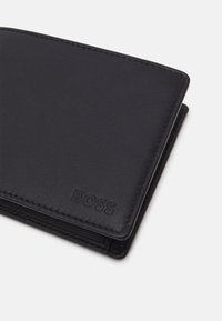 Portefeuille en cuir noir avec une texture lisse, présentant des coutures discrètes et un logo de marque en relief. Compact, de forme rectangulaire.