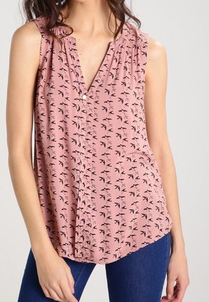 Mouwloze roze blouse met zwart vogeltjespatroon, V-hals, knopen aan de voorkant, gedragen met blauwe jeans door een persoon met donker haar.