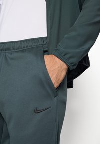 Zelen trenirni komplet Nike z jakno na zip in hlačami z elastičnim pasom. Ima majhen črn logotip ter gladko, teksturirano blago.