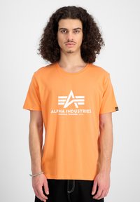 Kurze orange Baumwoll-T-Shirt mit weißem "Alpha Industries"-Logo und Schriftzug. Gerader Schnitt, lässige Passform.