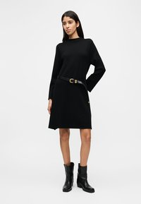 Γυναίκα με μαύρο φόρεμα μακριάς μανίκας, μαύρη ζώνη και μαύρες μποτάκια αστραγάλου, στέκεται με το ένα χέρι στη μέση της μπροστά σε απλό φόντο.