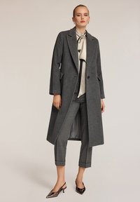 Cappotto di lana grigio con rever a punta, tasche laterali e chiusura con un singolo bottone, indossato sopra pantaloni a quadri grigi e scarpe nere con punta.
