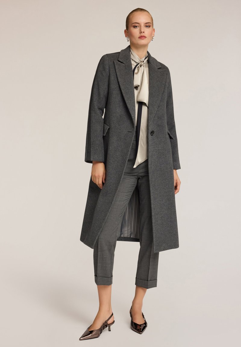 Cappotto di lana grigio con rever a punta, tasche laterali e chiusura con un singolo bottone, indossato sopra pantaloni a quadri grigi e scarpe nere con punta.