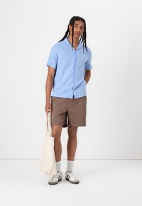 Chemise à manches courtes bleu clair avec des boutons, associée à un short marron. Le mannequin tient un sac fourre-tout beige, portant des baskets grises et blanches avec des chaussettes blanches.